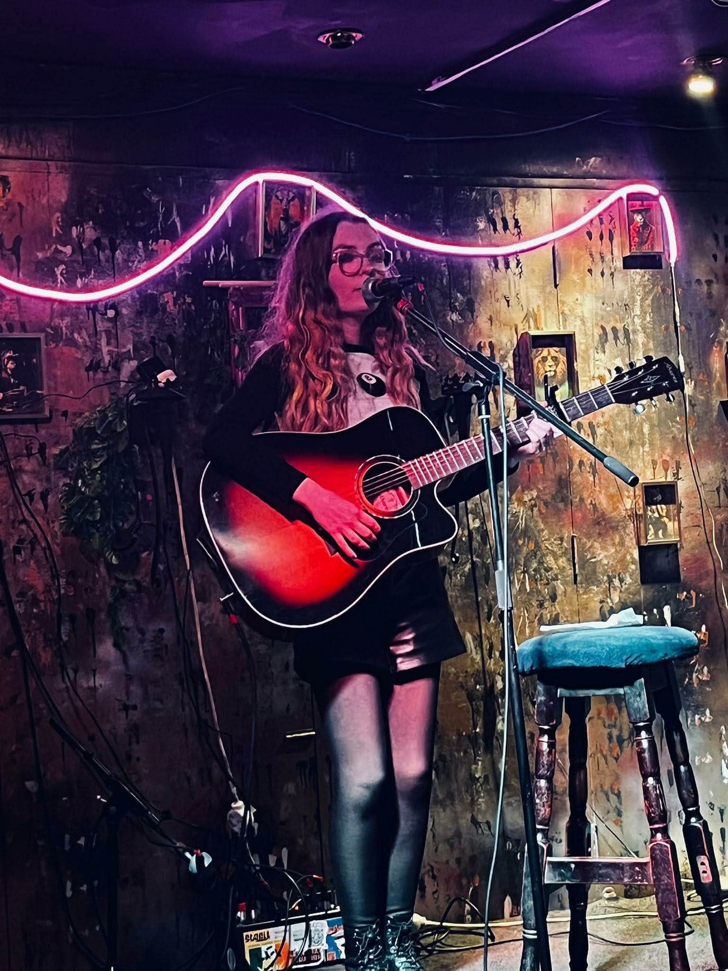Aimée O'Brien playing acoustic guitar on a small stage under a pink neon light