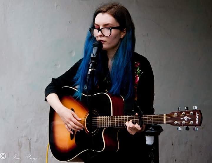 Aimée with blue hair, performing against a grey wall