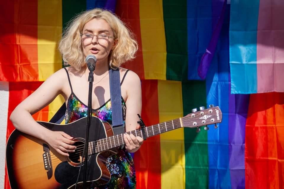 Aimée playing in front of a rainbow flag in the sun